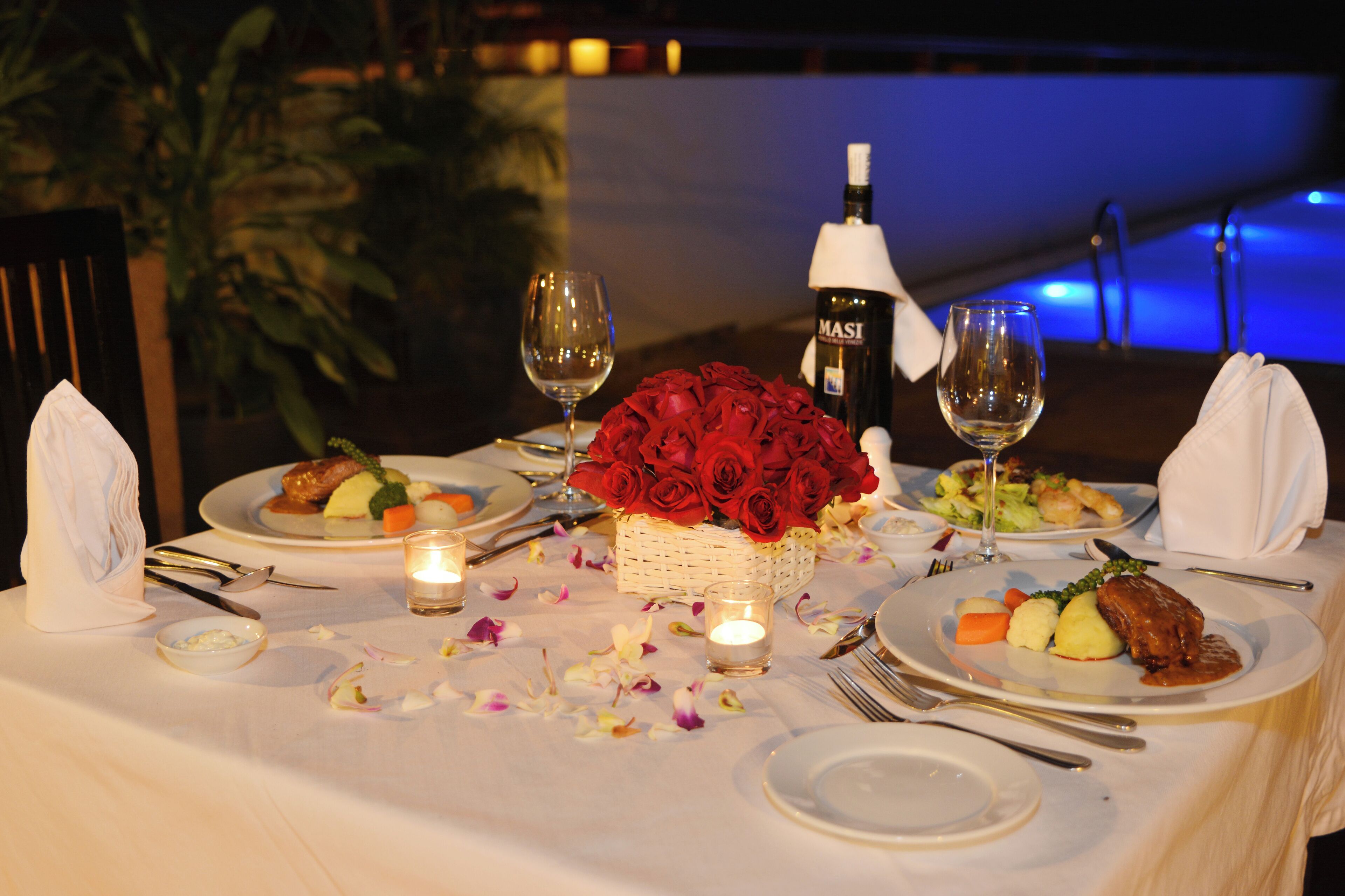 A table set for a romantic dinner with rose petals, candles, and wine.