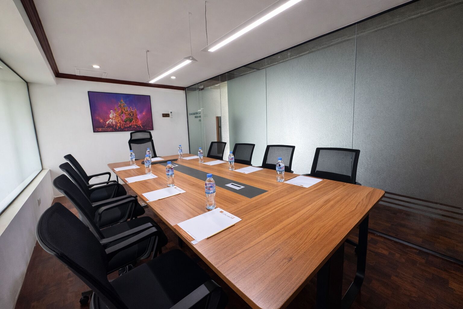 A conference room with a long wooden table, black chairs, water bottles, and a painting on the wall.