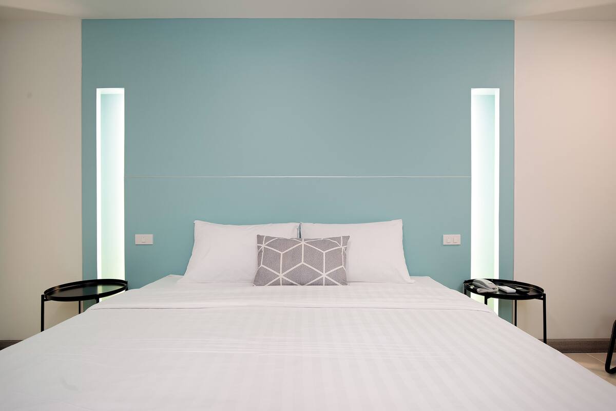 Een bed met witte beddengoed, een kussen met een geometrisch patroon, en twee zwarte nachtkastjes met een telefoon en een afstandsbediening erop.