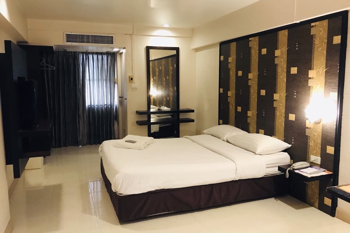 Una habitación de hotel con una cama, una mesita de noche con un teléfono y papeles, un espejo, un armario y una rejilla en el techo.