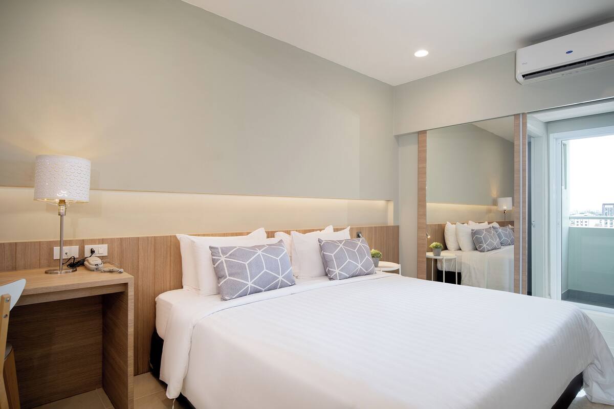 Een moderne hotelkamer met een groot bed, nachtkastje, lamp en uitzicht op de stad door een groot raam.