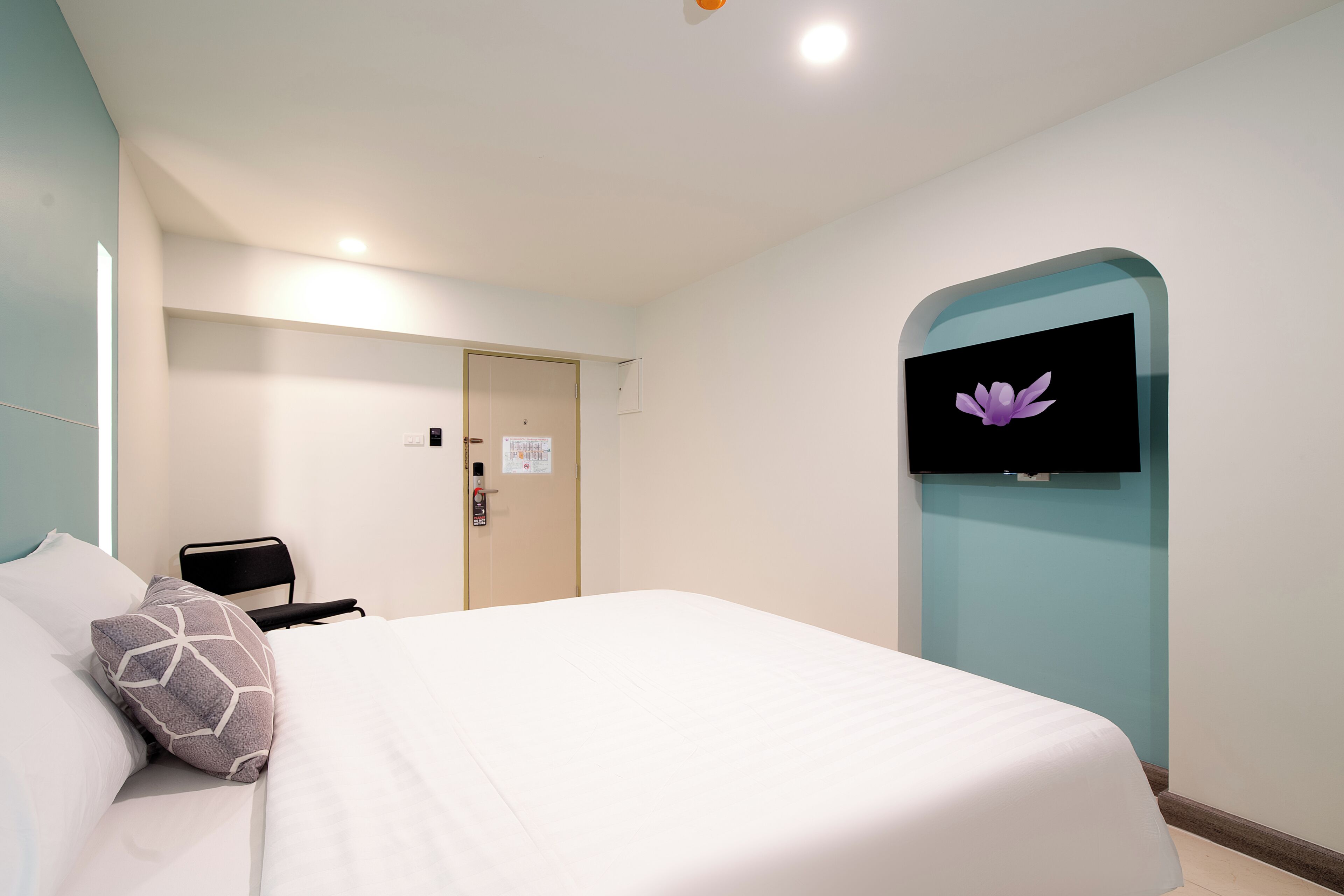 A hotel room with a bed, a TV mounted on the wall, a door, and a light fixture.