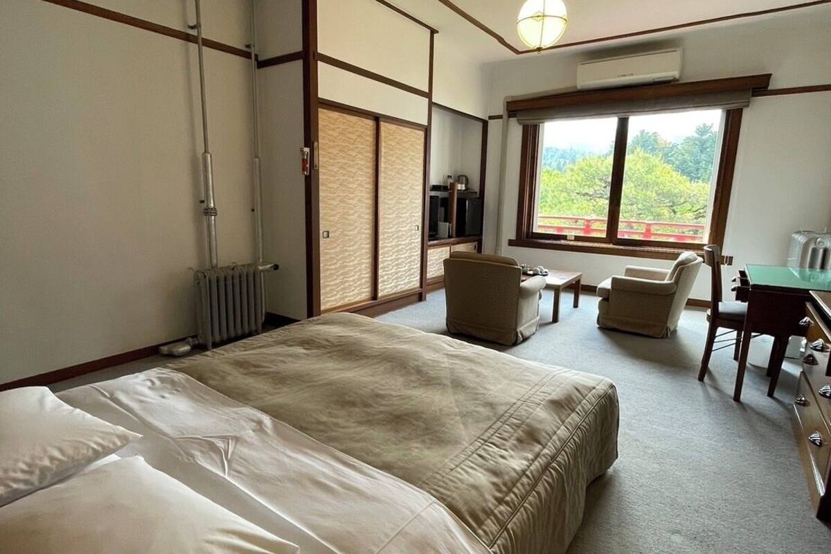 A hotel room with a large bed, a desk with a chair, a radiator, a television, and a window with a view of greenery.