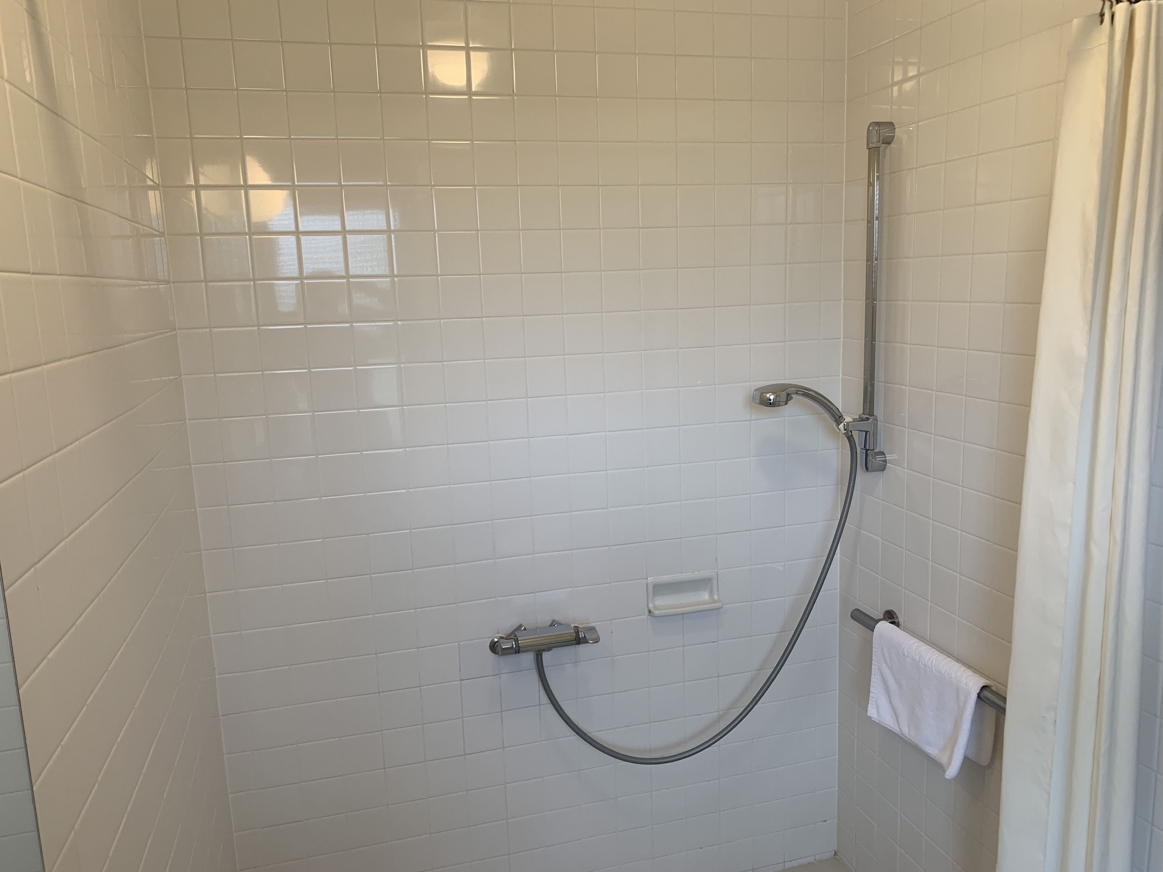 Área de duche com paredes em azulejo branco, chuveiro, mangueira de duche e toalheiro com uma toalha.