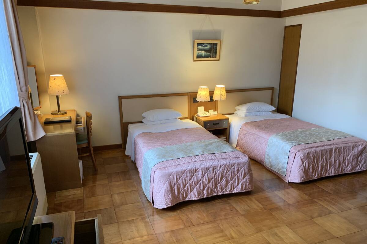 A hotel room with two beds, a desk with a lamp, a chair, and a wooden floor.