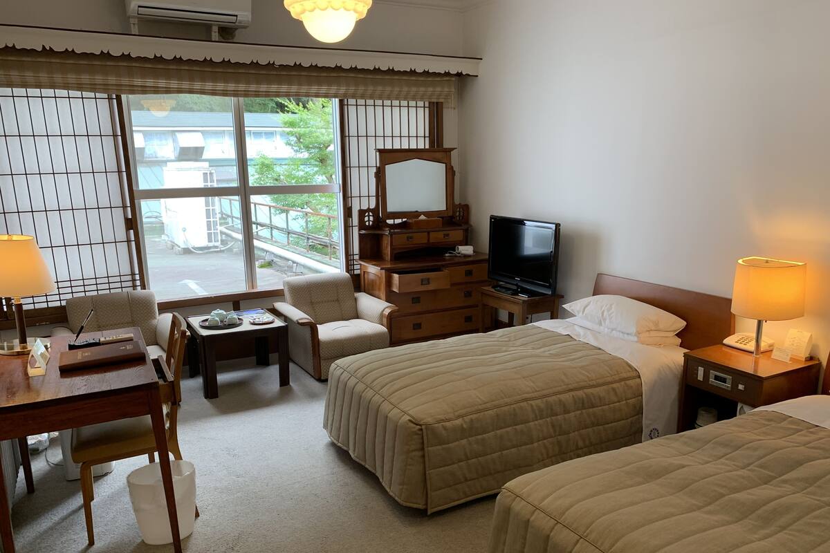 A hotel room with two beds, a desk with a chair, a television, and a window with a view of buildings and trees.