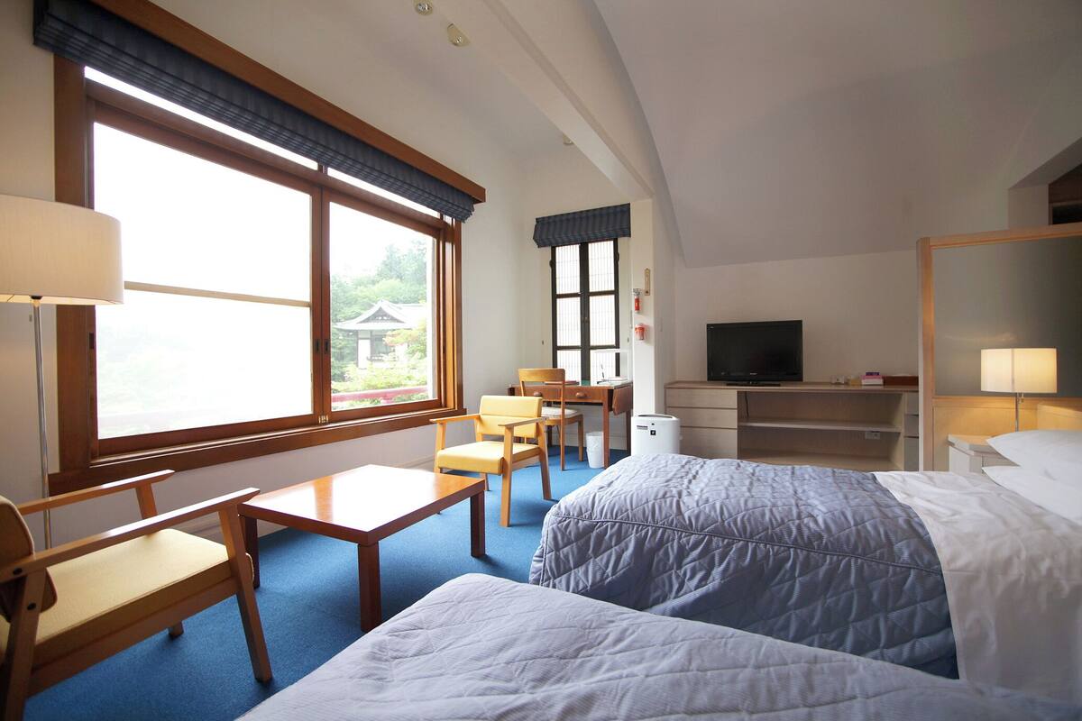 A hotel room with two beds, a desk, a chair, a TV, and large windows.