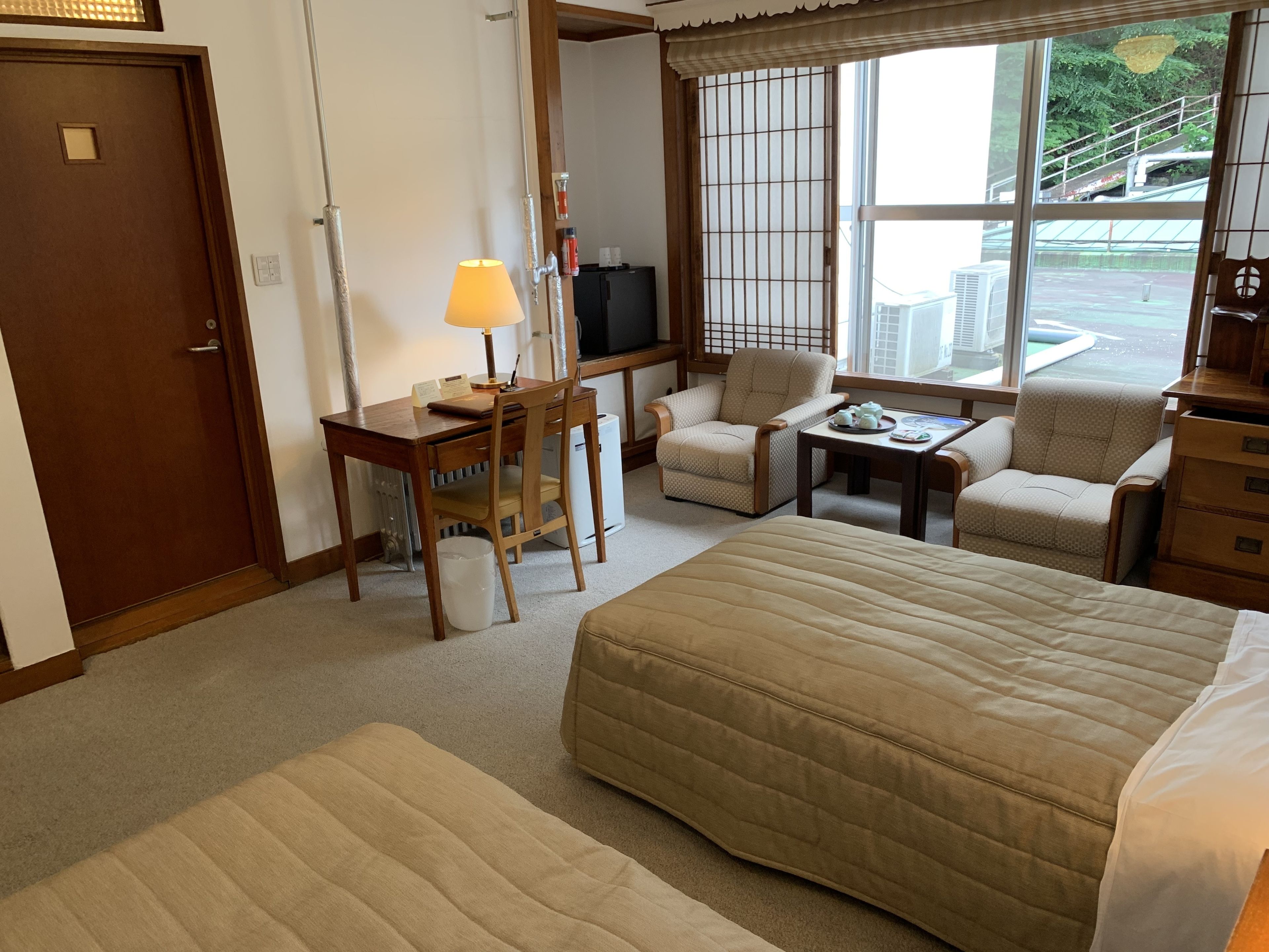 A hotel room with a large bed, two armchairs, a small table, a desk with a lamp, a TV, and a window with a view of the outside.