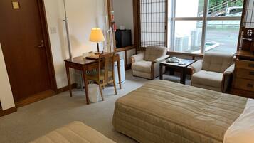 A hotel room with a large bed, two armchairs, a small table, a desk with a lamp, a TV, and a window with a view of the outside.