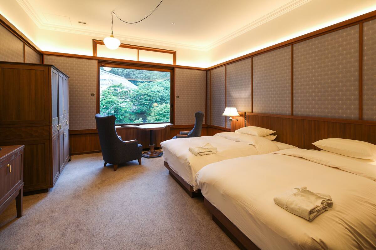A hotel room with two beds, a desk, a chair, and a large window with a view of greenery.