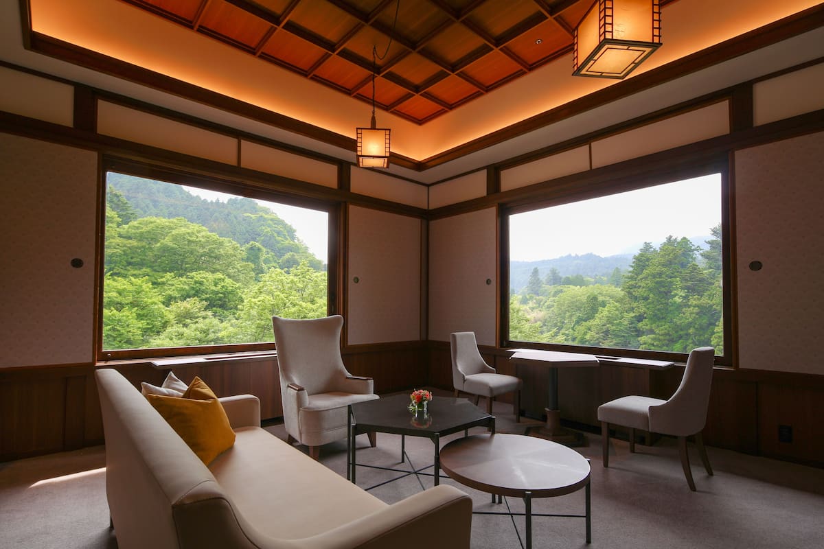 A room with a sofa, two armchairs, a coffee table, and a desk with a view of a forest through large windows.