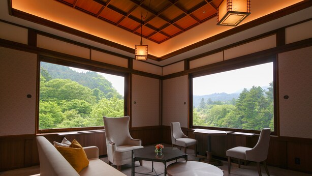 Habitación con sofá, dos sillones, una mesa de centro y un escritorio, con vistas a un bosque a través de amplios ventanales.