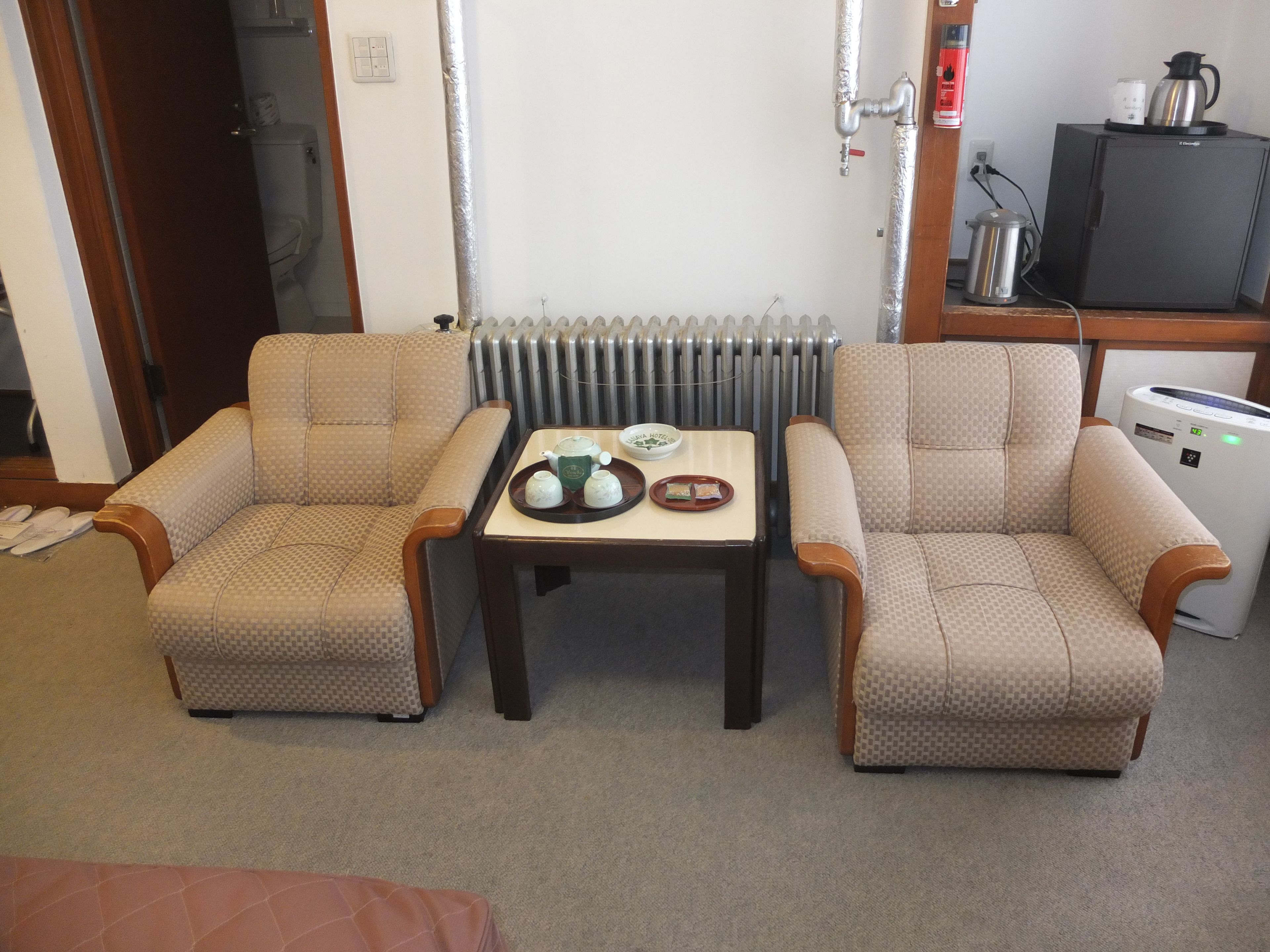 Ein kleines, möbliertes Zimmer mit zwei gestreiften Sesseln, einem kleinen Tisch mit einer Teekopfsammlung, einem Heizkörper, einer Kaffeemaschine und einem Fernseher.