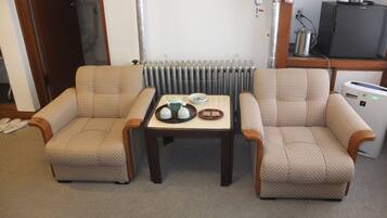 Ein kleines, möbliertes Zimmer mit zwei gestreiften Sesseln, einem kleinen Tisch mit einer Teekopfsammlung, einem Heizkörper, einer Kaffeemaschine und einem Fernseher.