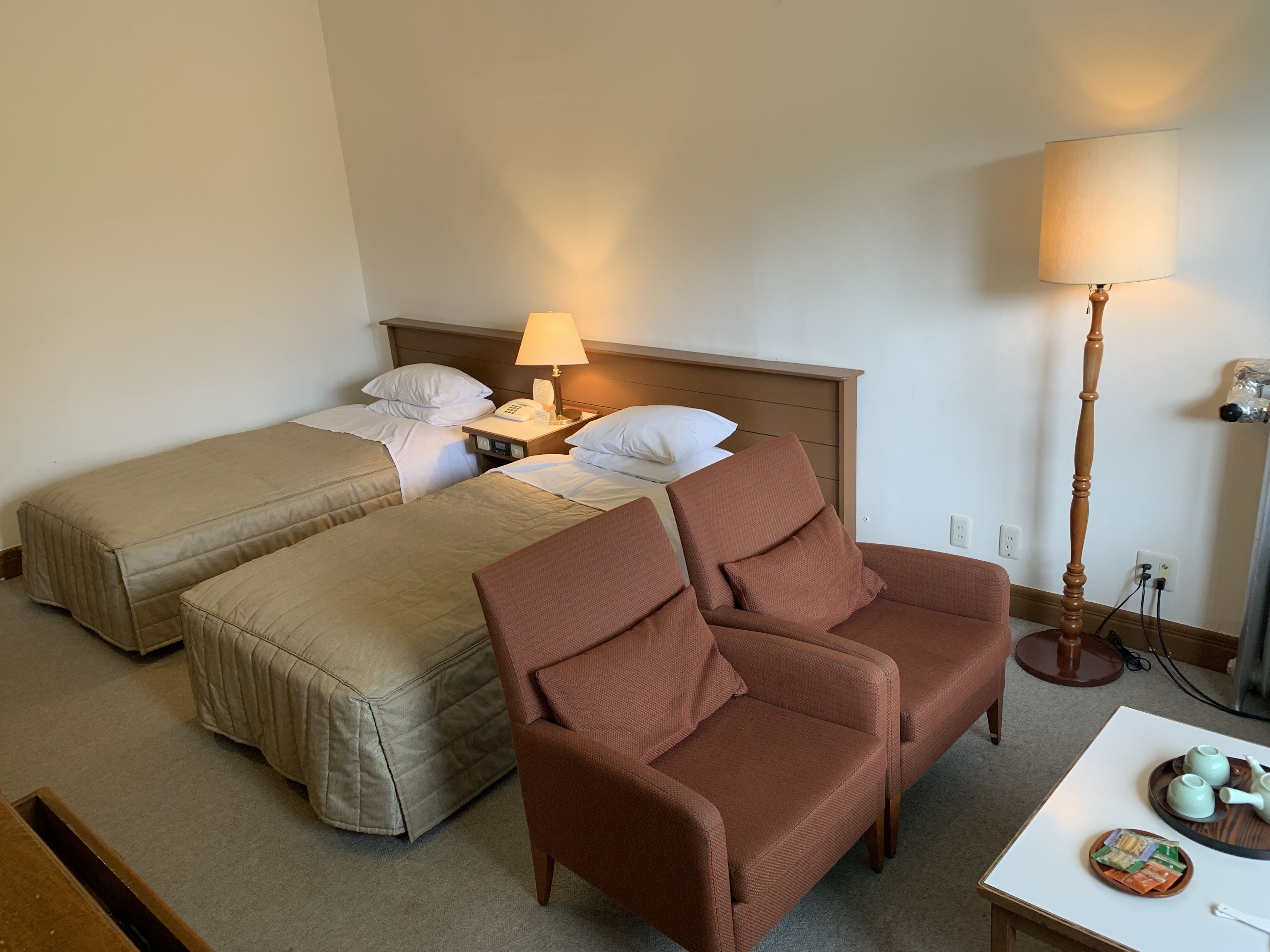 A hotel room with two single beds, a brown armchair, a small table with a tray of tea and snacks, and a bedside lamp.