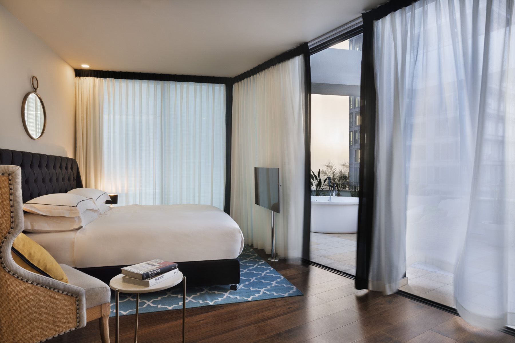 A modern hotel room with a large bed, a flat-screen TV, a small table with books, and a view through a glass door.