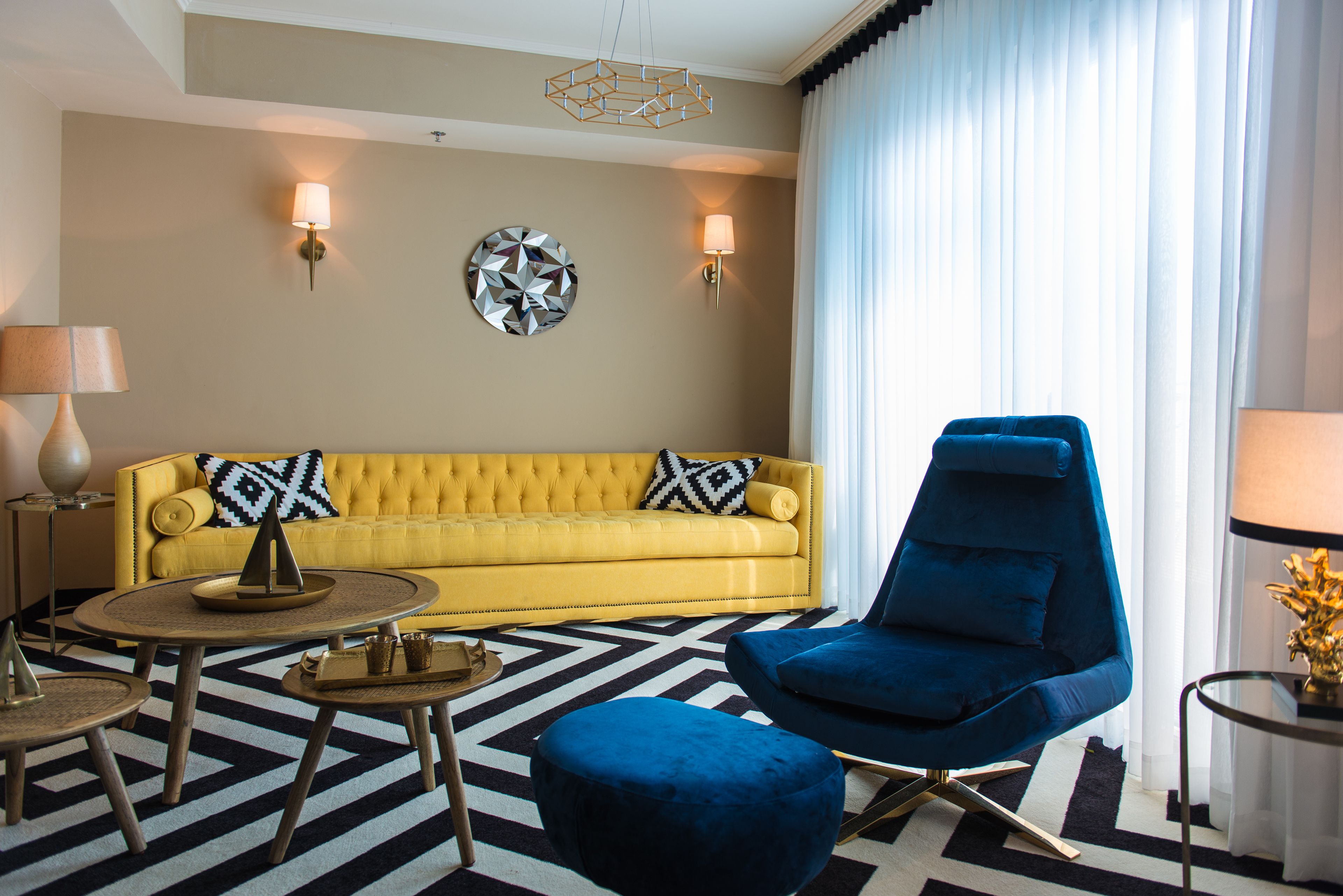 Un canapé jaune orné d’oreillers à motifs noirs et blancs, un fauteuil bleu, une table basse ronde et un élément de décoration murale aux motifs géométriques.