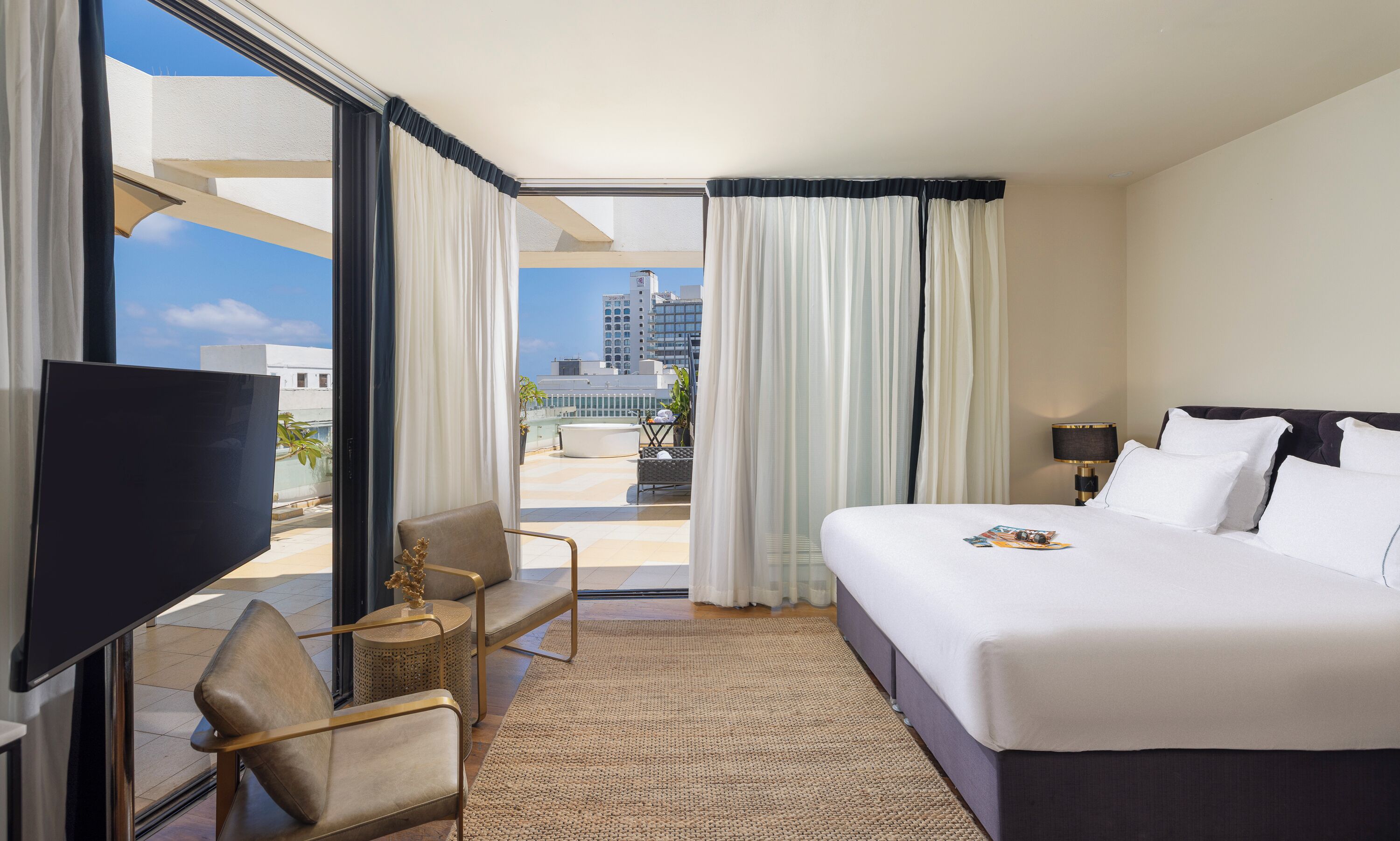 A hotel room with a bed, a TV, a chair, a small table, and a view of the cityscape.