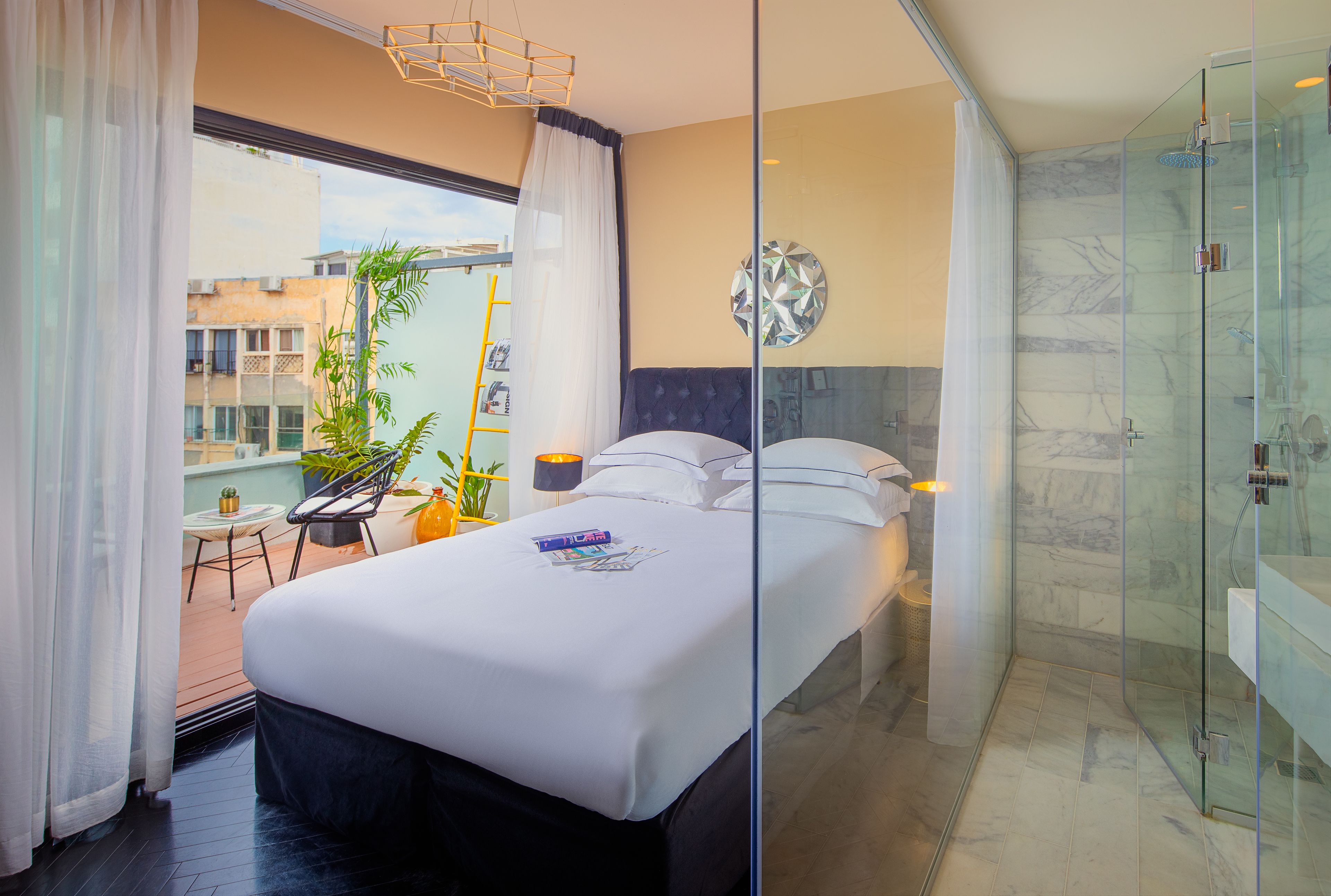 A modern hotel room with a large bed, a glass shower enclosure, a balcony with a chair and plants, and a city view.