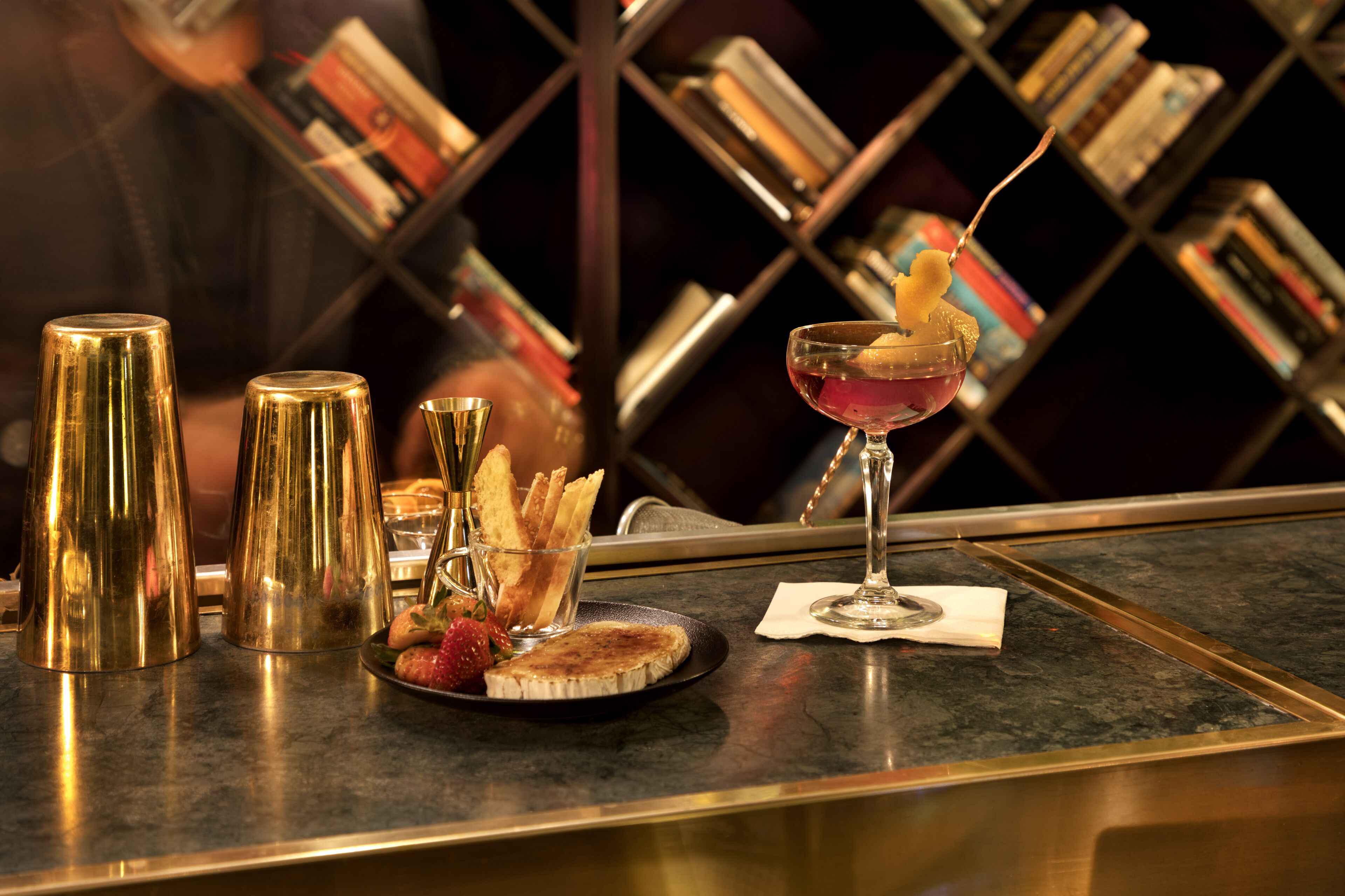 A bar setting with a martini glass, gold tumblers, and a plate with bread and strawberries.