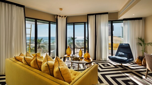 Un salon moderne avec un canapé jaune, un fauteuil bleu et un tapis à motifs. De grandes fenêtres offrent une vue sur la plage et les palmiers.