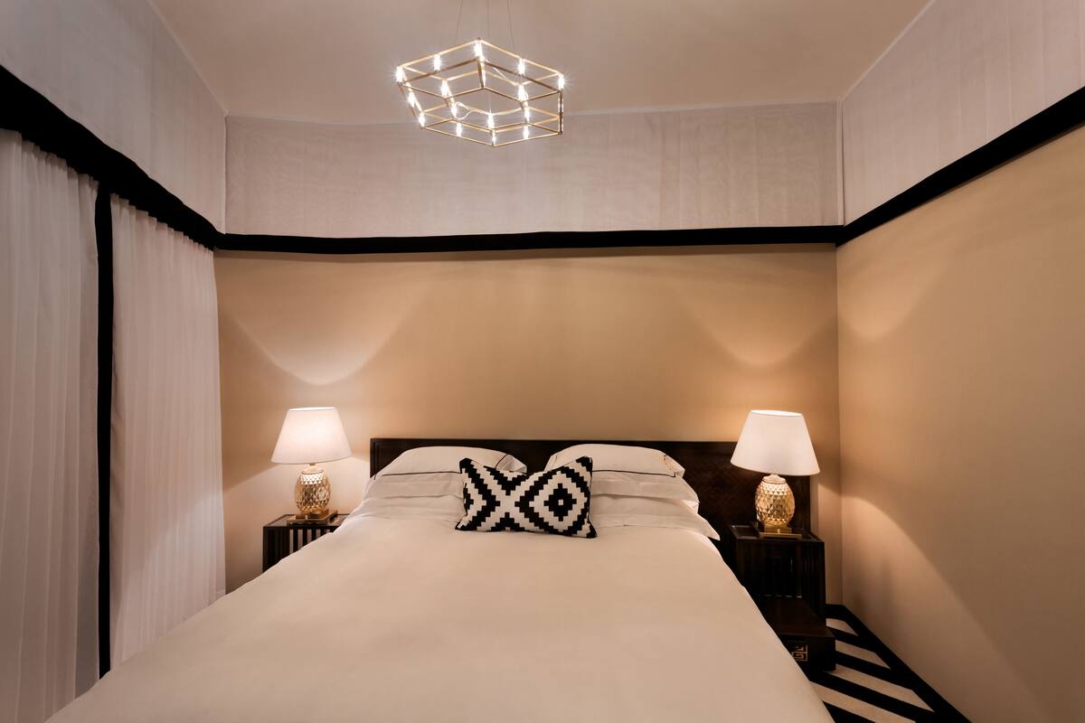 Une chambre d’hôtel avec un grand lit, deux tables de chevet avec des lampes, un luminaire au plafond au design géométrique et des rideaux légers.