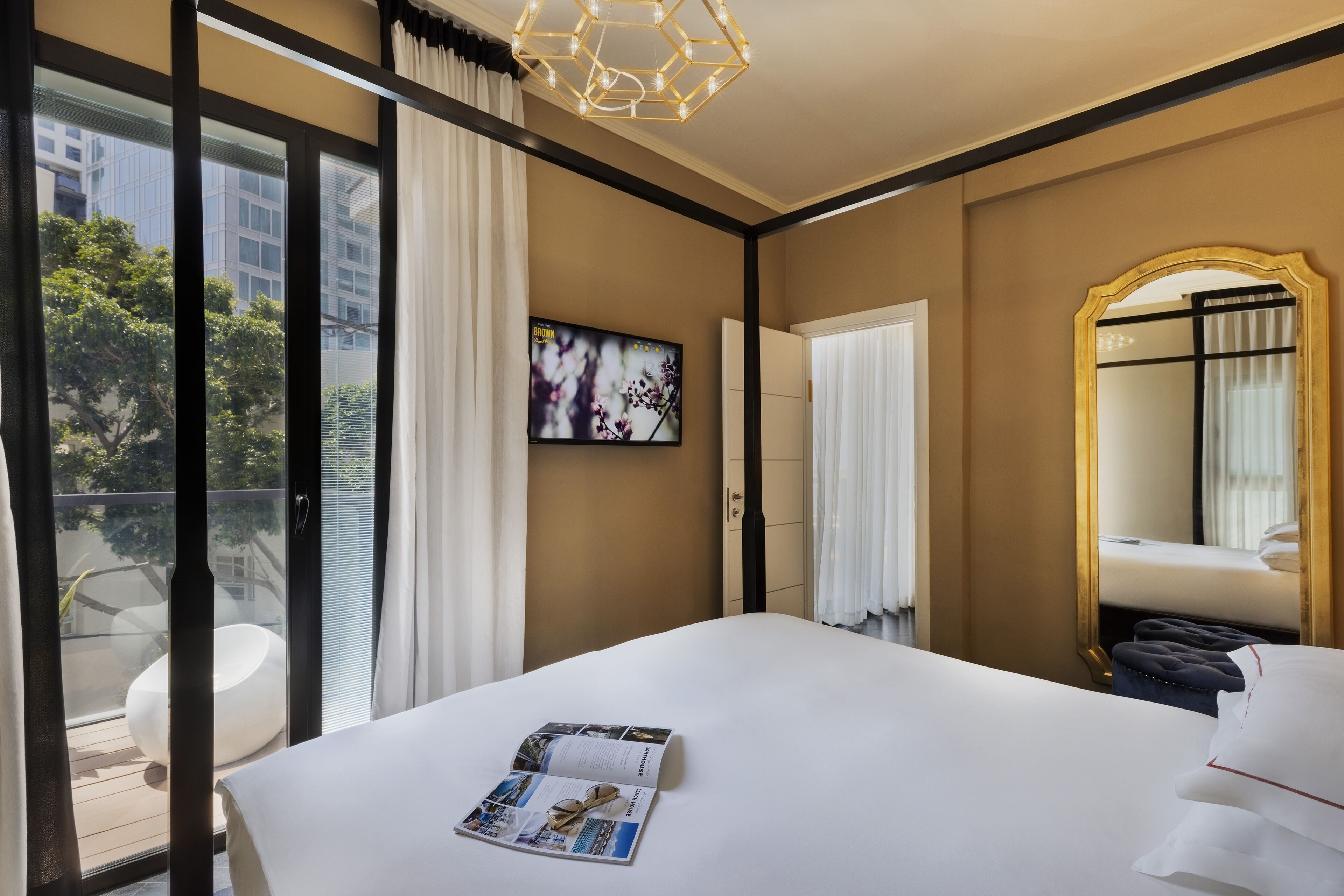 Habitación de hotel con una cama grande, un televisor en la pared, un balcón con una silla blanca y vistas a edificios.