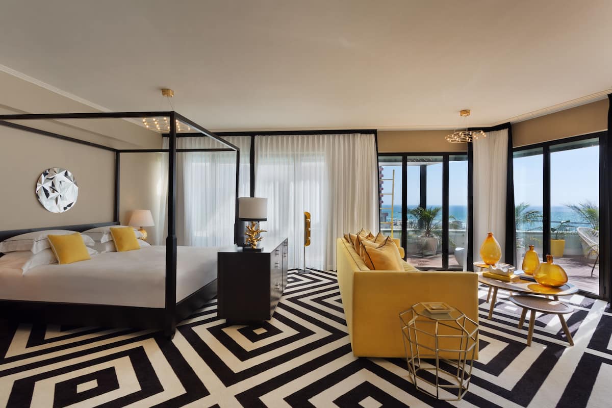 Une chambre d’hôtel moderne avec un lit à baldaquin, un canapé jaune, une table à manger agrémentée de vases jaunes et de grandes fenêtres offrant une vue sur l’océan.