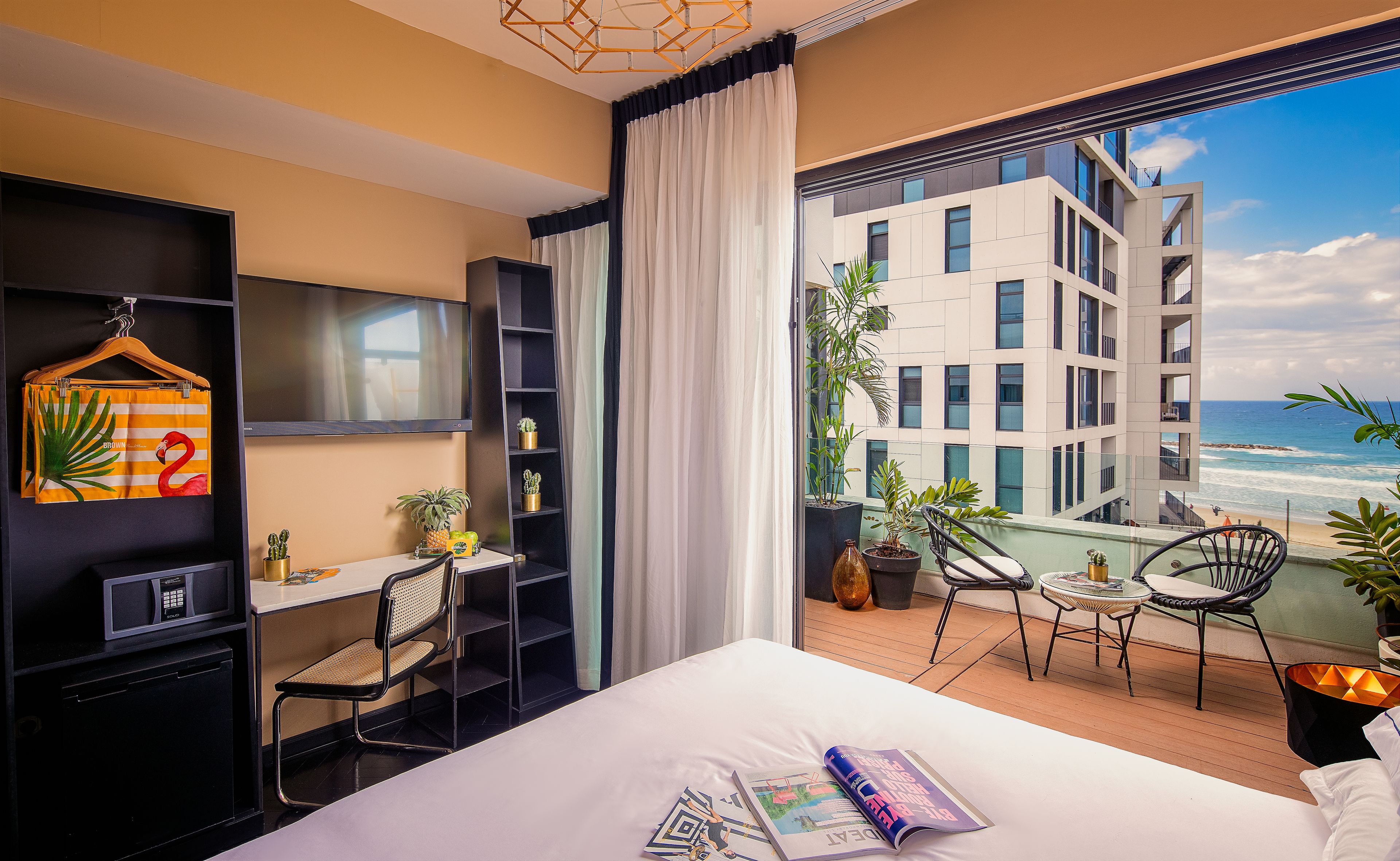 Ein modernes Hotelzimmer mit einem großen Bett, einem Schreibtisch mit Stuhl, einem Fernseher und einem Balkon mit Blick auf das Meer und ein Gebäude.