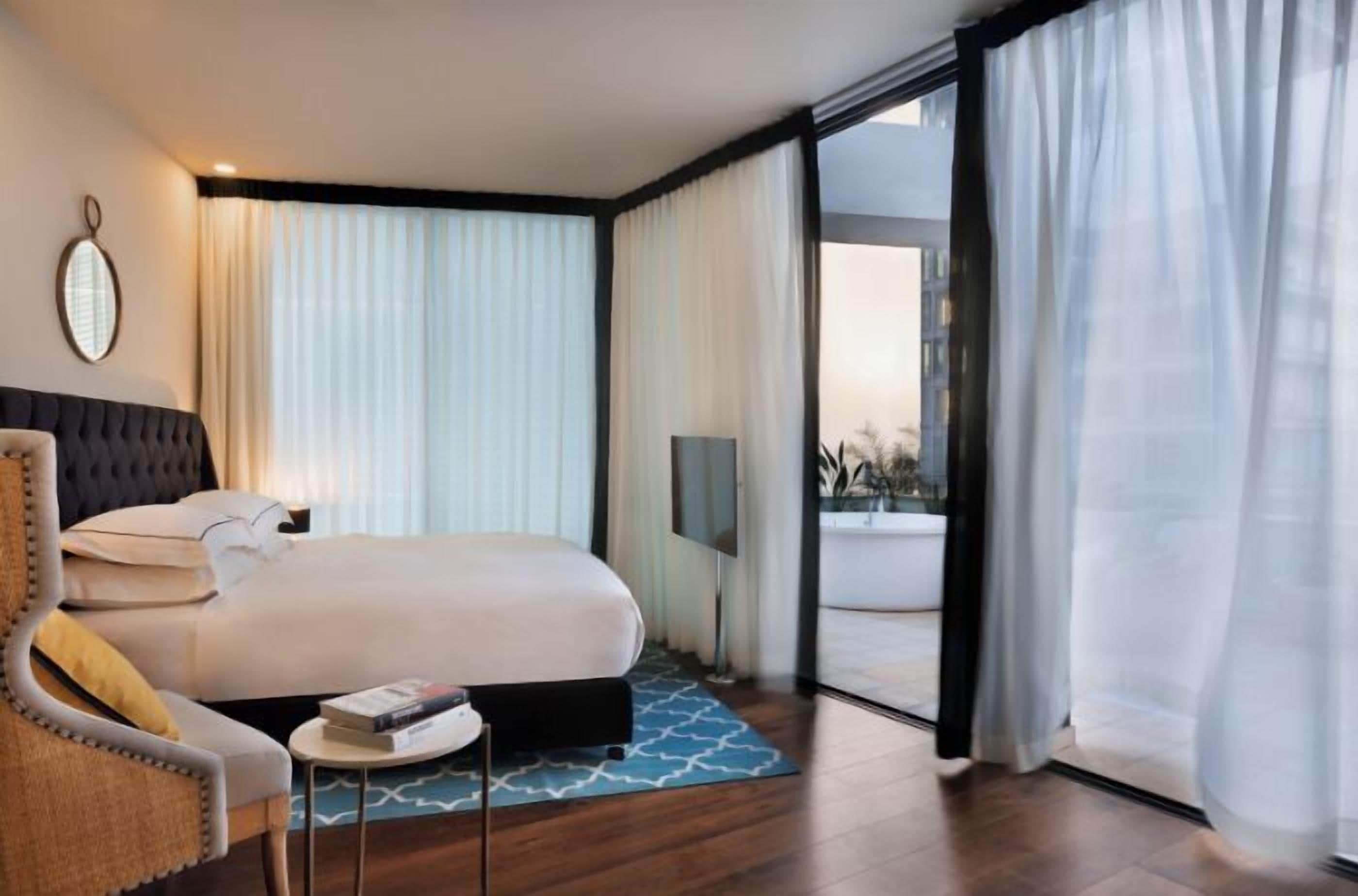 A modern hotel room with a large bed, a small side table, a chair, and a large window with curtains.
