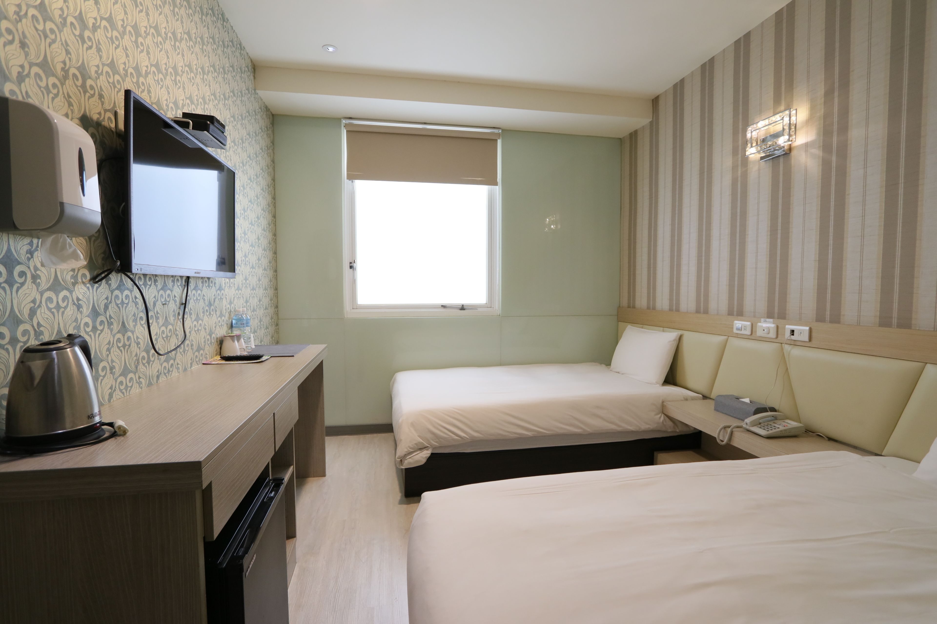 Habitación de hotel con dos camas, un escritorio con hervidor, televisor y ventana con persiana color crema.
