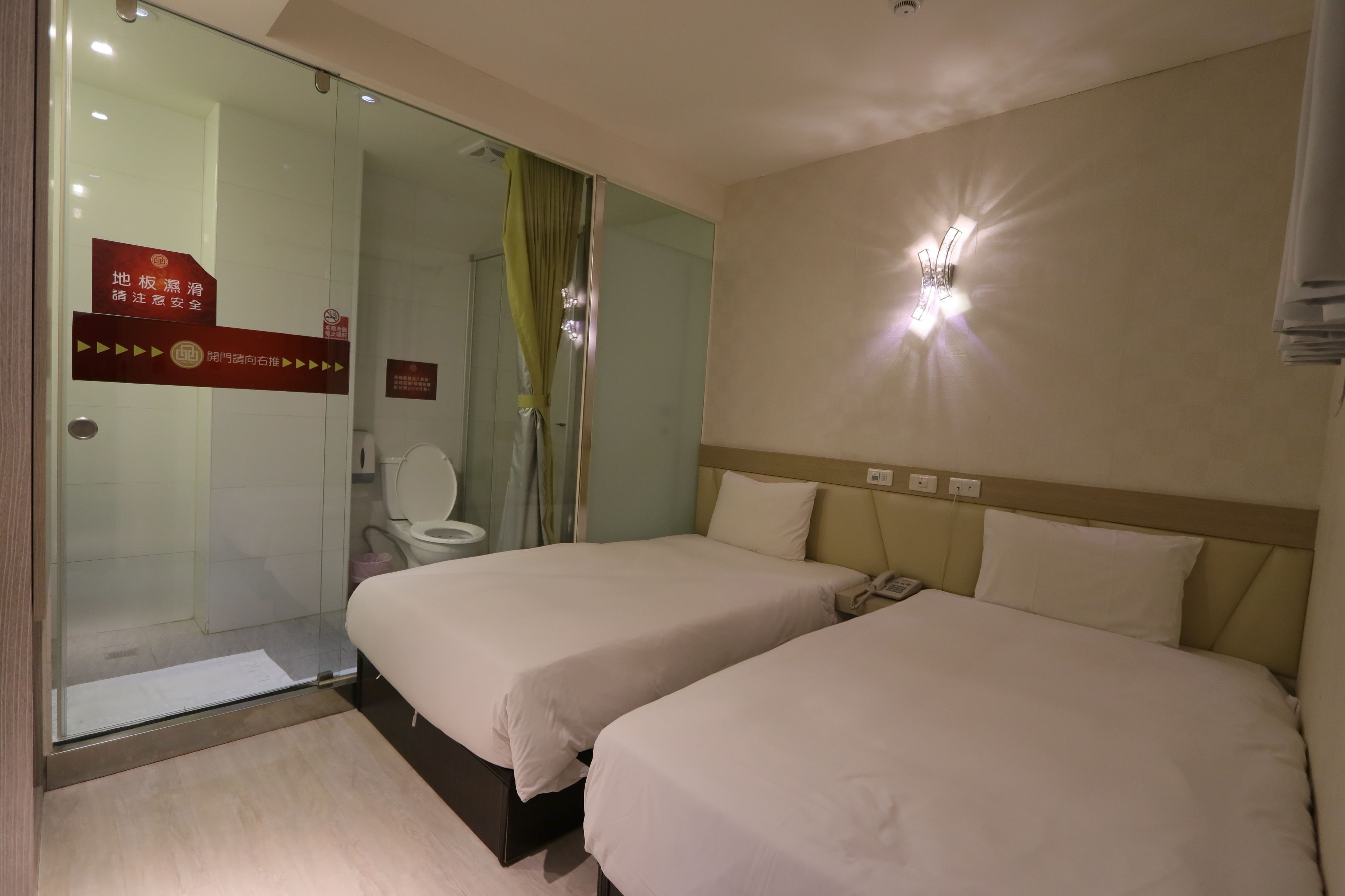 A hotel room with two beds, a shower area, and a toilet. There are signs on the door indicating the location and safety instructions.