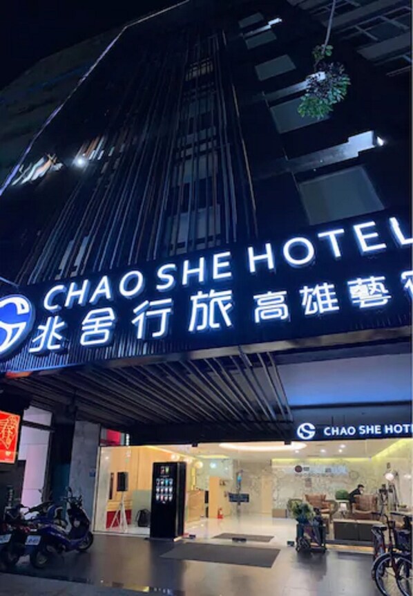 L’entrée d’un hôtel, avec une enseigne indiquant « CHAOSHE HOTEL » et une autre avec des caractères chinois. On peut voir des vélos garés à l’extérieur et une personne dans l’embrasure de la porte.