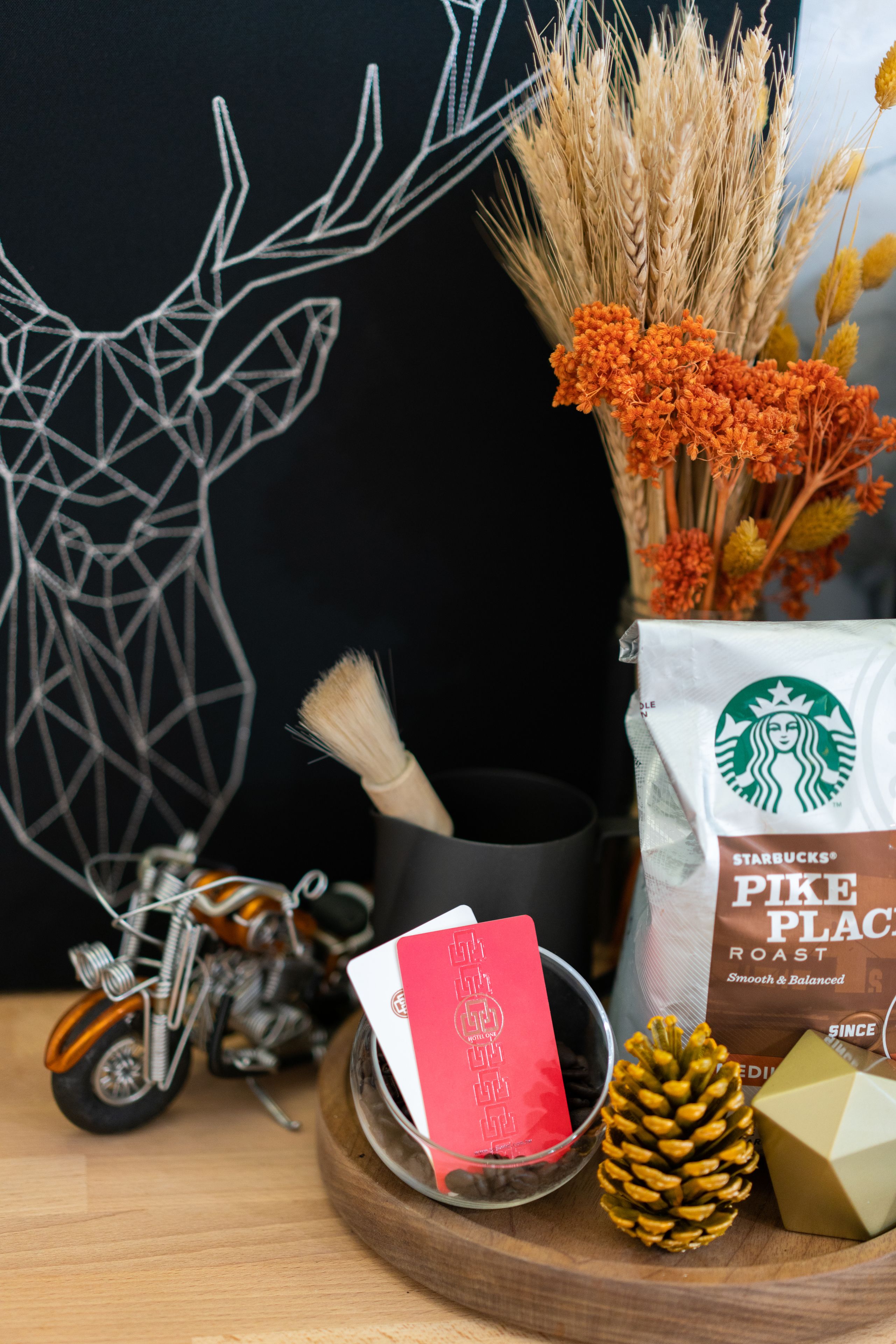 A wooden tray with a Starbucks Pike Place Roast bag, a red card, a glass bowl, a pine cone, and a decorative metal sculpture.