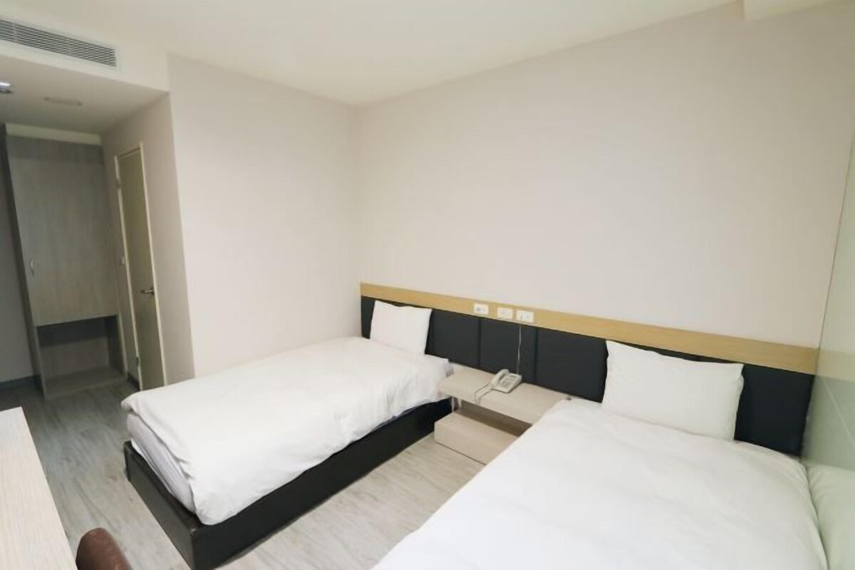 Una habitación de hotel con dos camas, una mesita de noche con teléfono y una puerta que da a otra habitación.