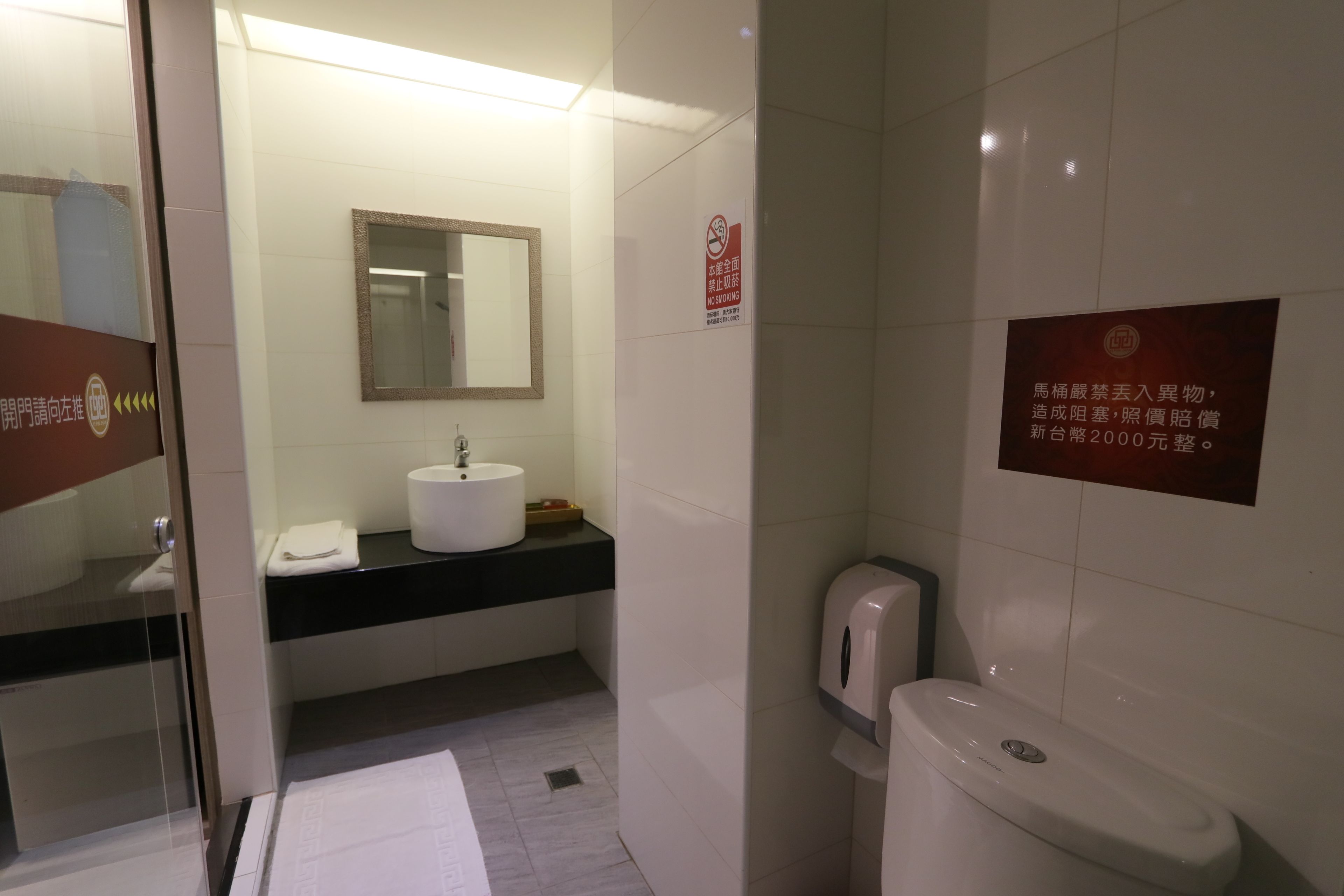 A modern bathroom with a sink, mirror, and toilet. There is a no-smoking sign on the wall and a sign indicating a fine for smoking.