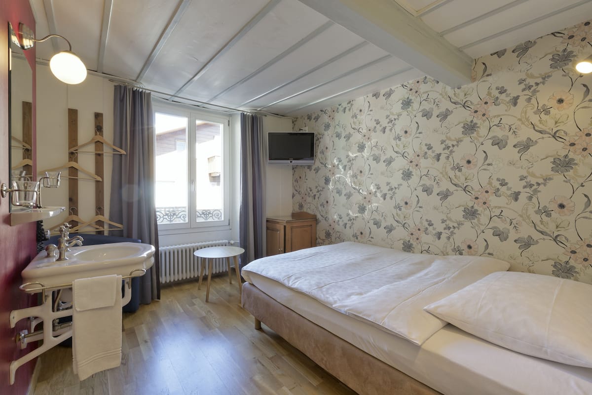 A bedroom with a bed, a bedside table, a TV, a window, and a bathroom with a sink and mirror.