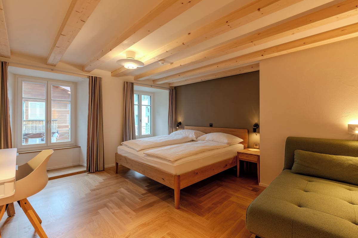 A bedroom with a wooden bed, a green armchair, a desk, and a window with curtains.