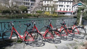 Une rangée de vélos rouges est stationnée le long d’une promenade au bord de la rivière, avec des bâtiments en arrière-plan.
