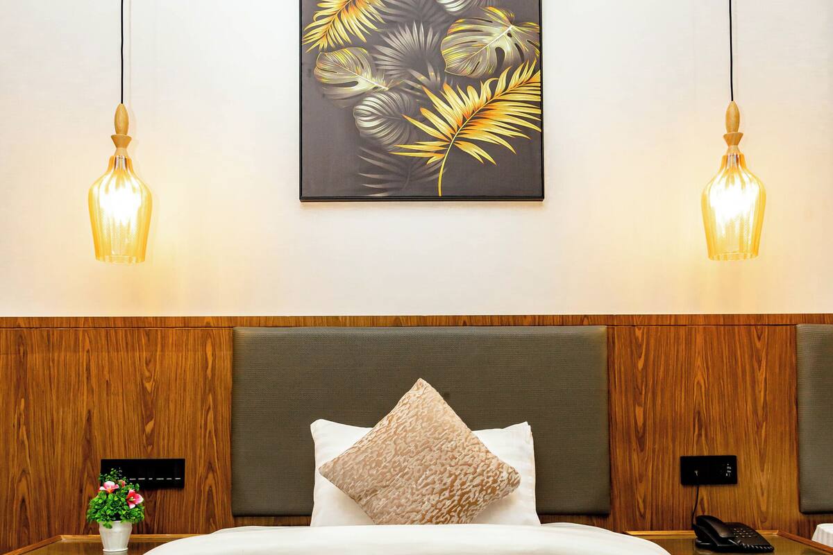 A hotel room with a bed, two bedside tables, a nightstand, a wall-mounted painting, and two hanging lamps.