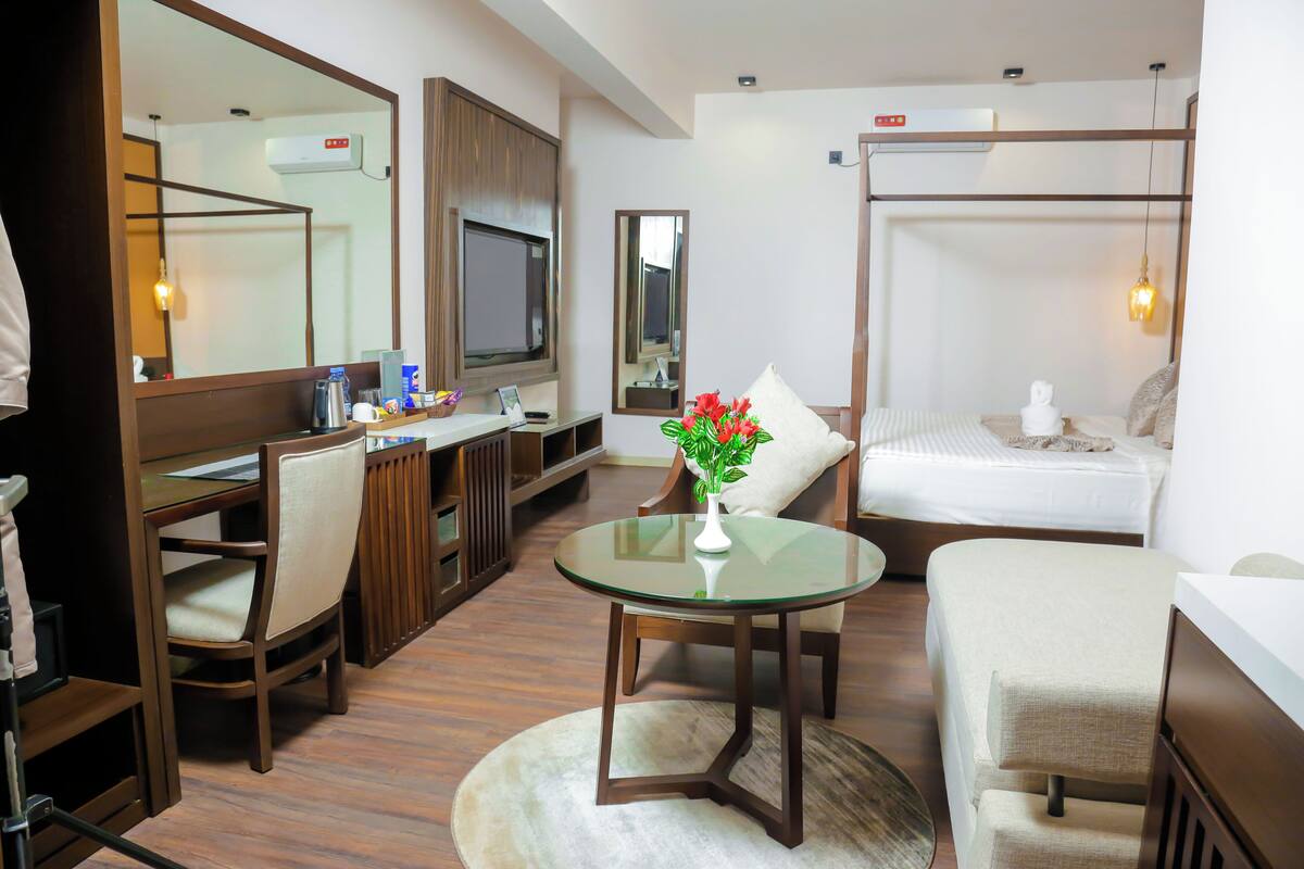 A hotel room with a bed, a desk with a chair, a round table with a vase of flowers, and a large mirror.