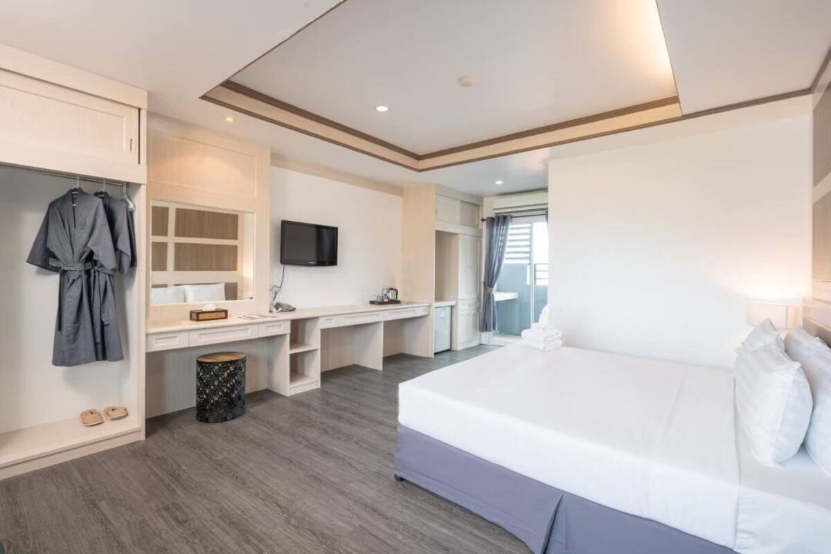 A modern hotel room with a large bed, a desk with a flat-screen TV, a wardrobe, and a small trash bin.
