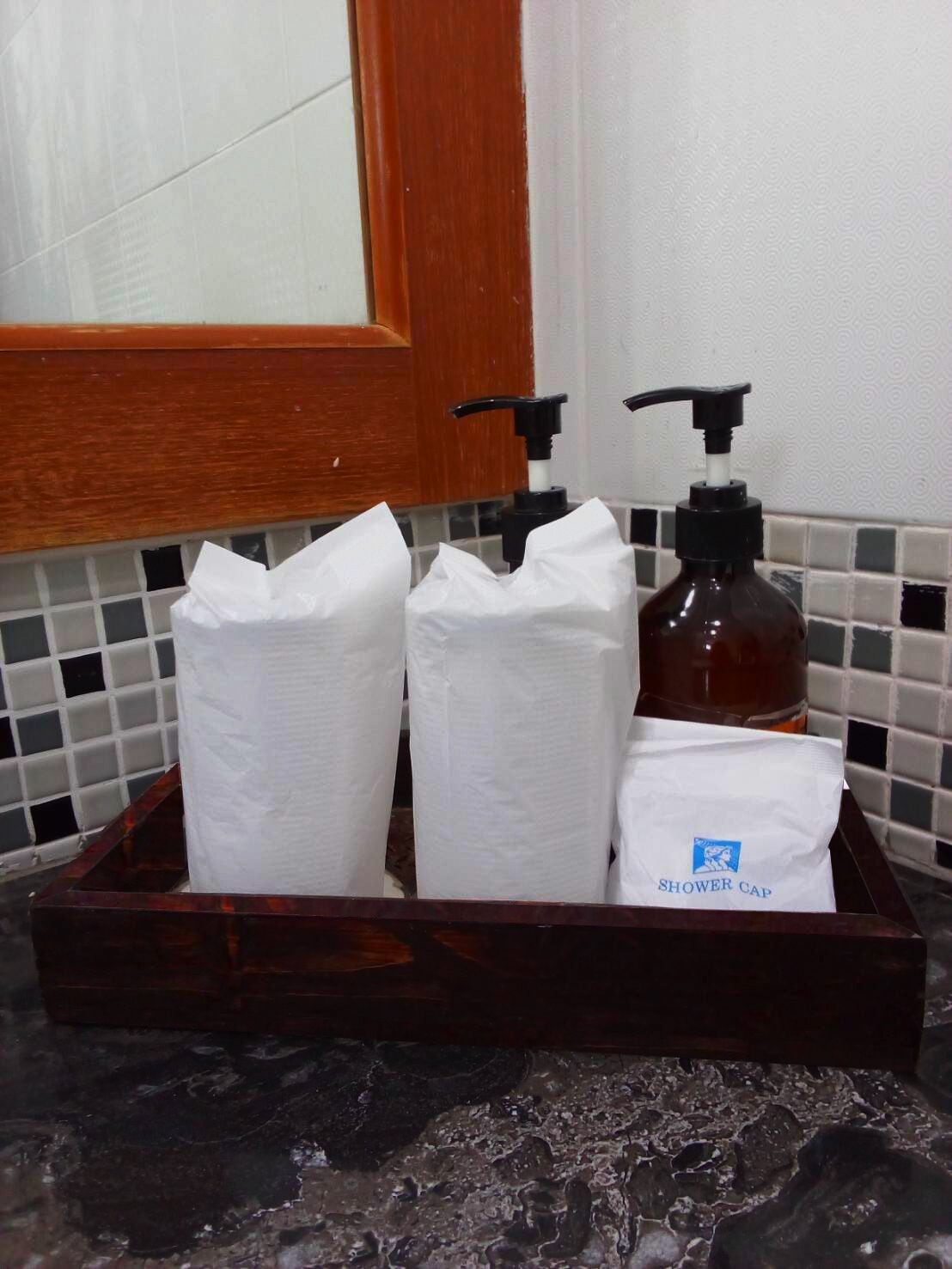 Un baño con una bandeja de madera que contiene una botella de jabón líquido, una toalla de papel y un gorro de ducha.