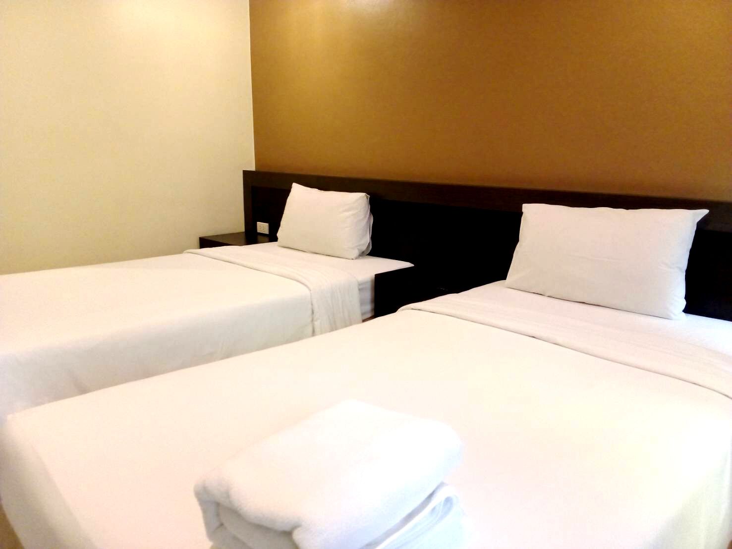 A hotel room with two single beds, each with a white bedspread and pillow, and a neatly folded white towel on the bed.