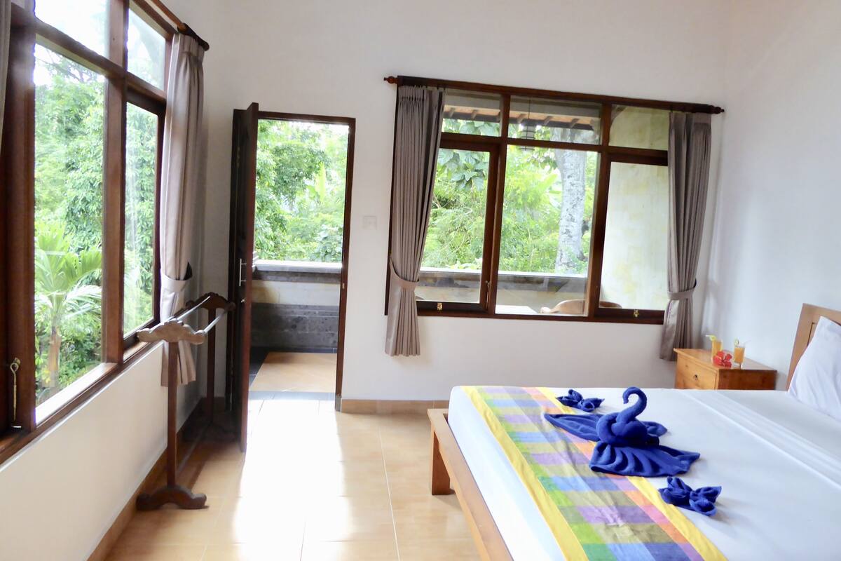 A bedroom with a bed, two windows, a wooden door, and a view of greenery outside.