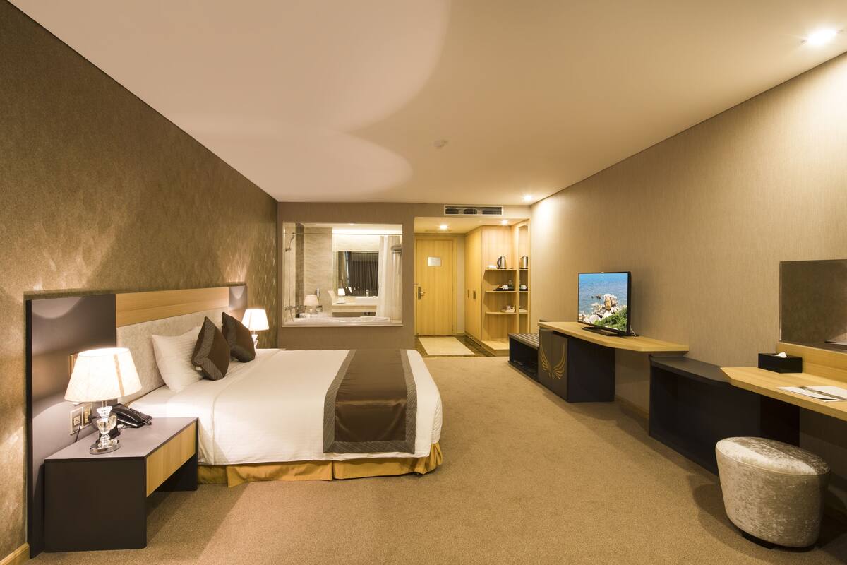 A hotel room with a bed, bedside table with a lamp, a TV, a desk, and a bathroom visible through an open door.