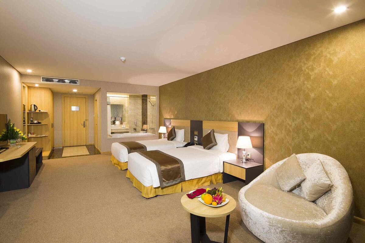 A hotel room with two beds, a sitting area with a chair, a small table with fruit, and a desk with a lamp.