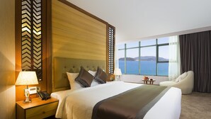 A hotel room with a large bed, a nightstand with a lamp, a chair, and a view of the sea. - Muong Thanh Luxury Nha Trang Hotel (Nha Trang)