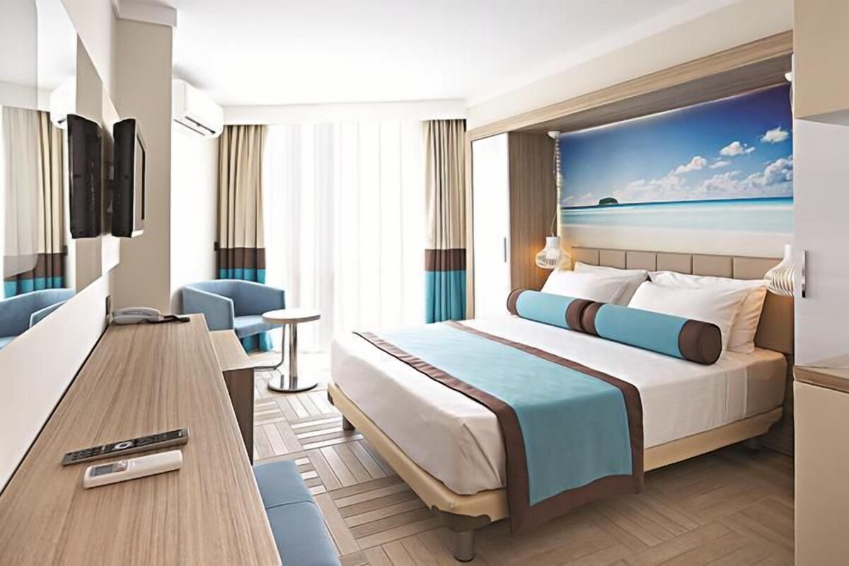 A hotel room with a bed, a desk, a chair, a TV, and a window with a view of the sea.