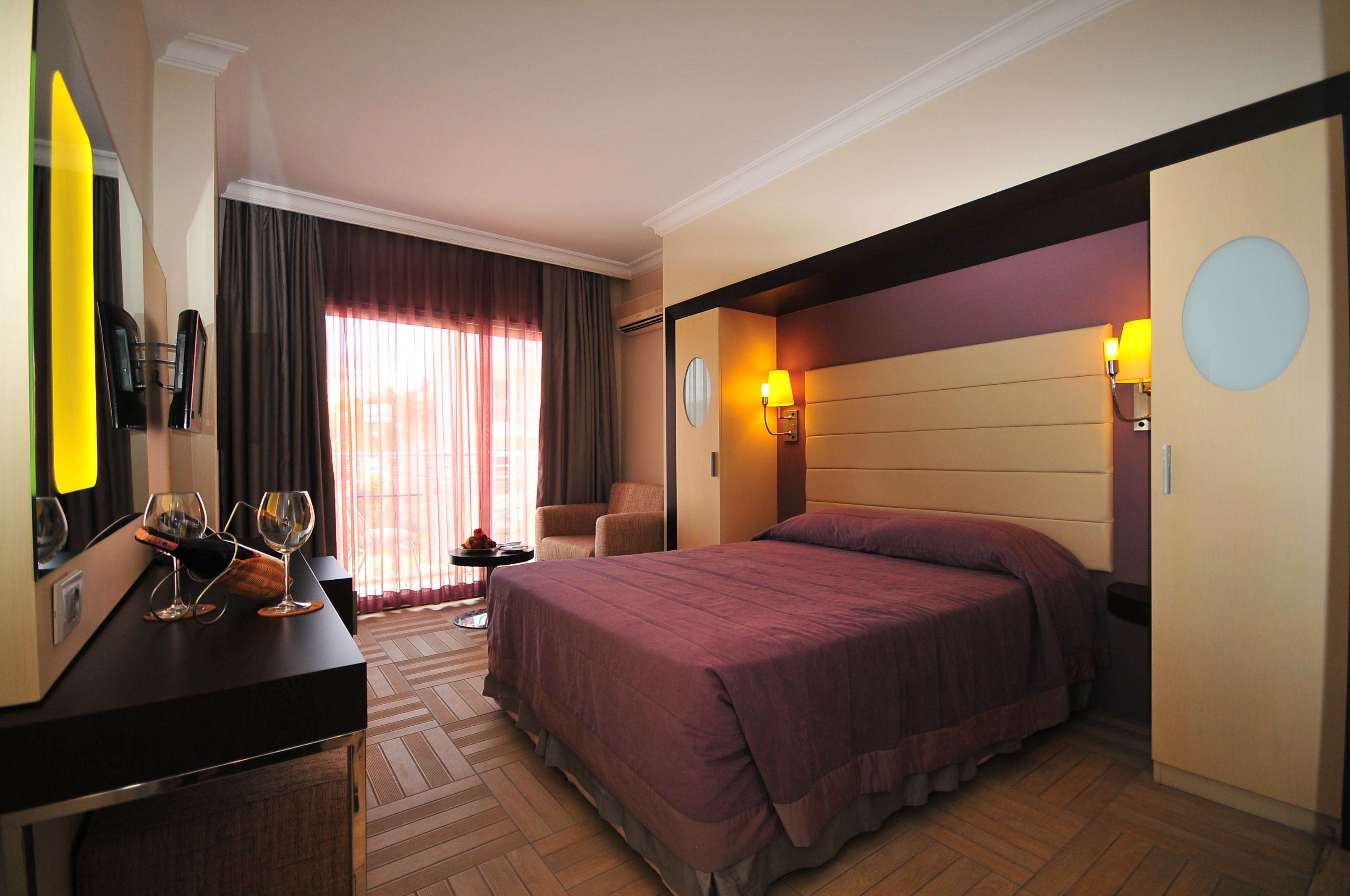 Hotelkamer met een groot bed, een bureau met stoel, een tafeltje met lamp en een raam met gordijnen.